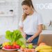 Woman in white t-shirt preparing fresh vegetables in a bright kitchen with a laptop.